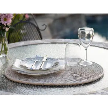 Round woven gray rattan placemat with white scalloped plate, silverware, napkin and clear wine glass on outdoor table