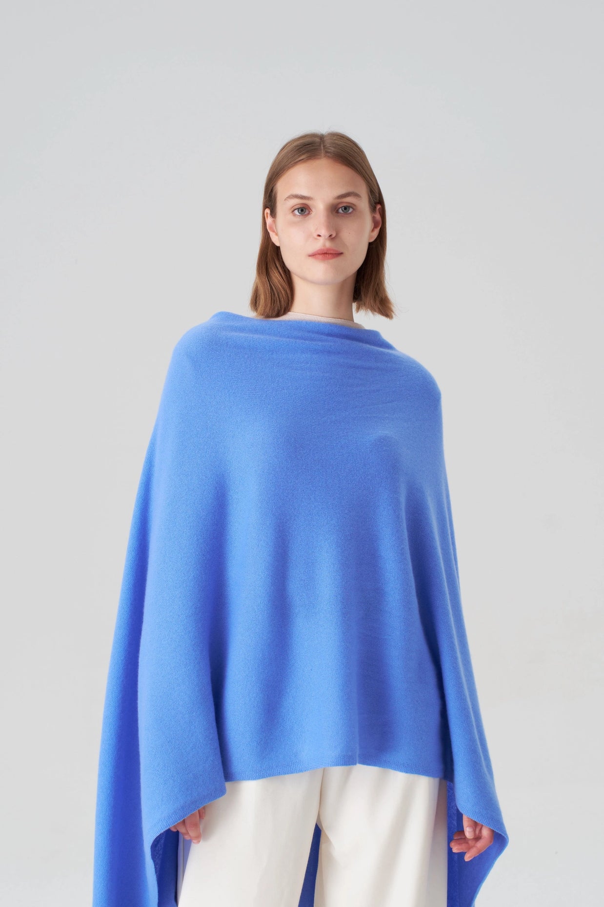 Periwinkle blue cashmere poncho with asymmetric drape and ribbed knit texture worn over cream wide-leg trousers