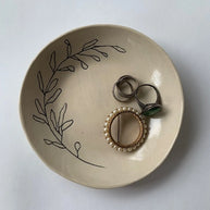 Cream ceramic ring dish with hand-drawn botanical leaf design holding silver and pearl-beaded rings on white surface