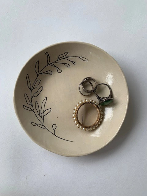 Cream ceramic ring dish with hand-drawn botanical leaf design holding silver and pearl-beaded rings on white surface