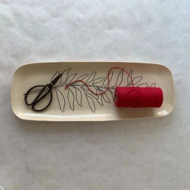 Cream ceramic tray with black botanical leaf design holding small black scissors and bright pink thread spool