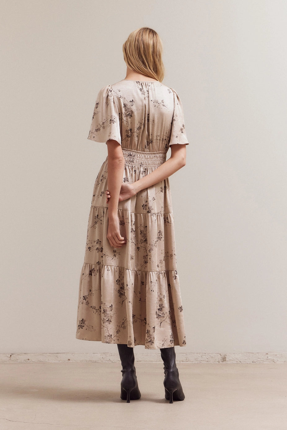 Beige tiered midi dress with brown floral print, smocked waist, and flutter sleeves shown from back view with black boots
