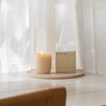 Cream-colored soy wax candle with lit flame on wooden tray beside beige Boheme box against white sheer curtain backdrop.