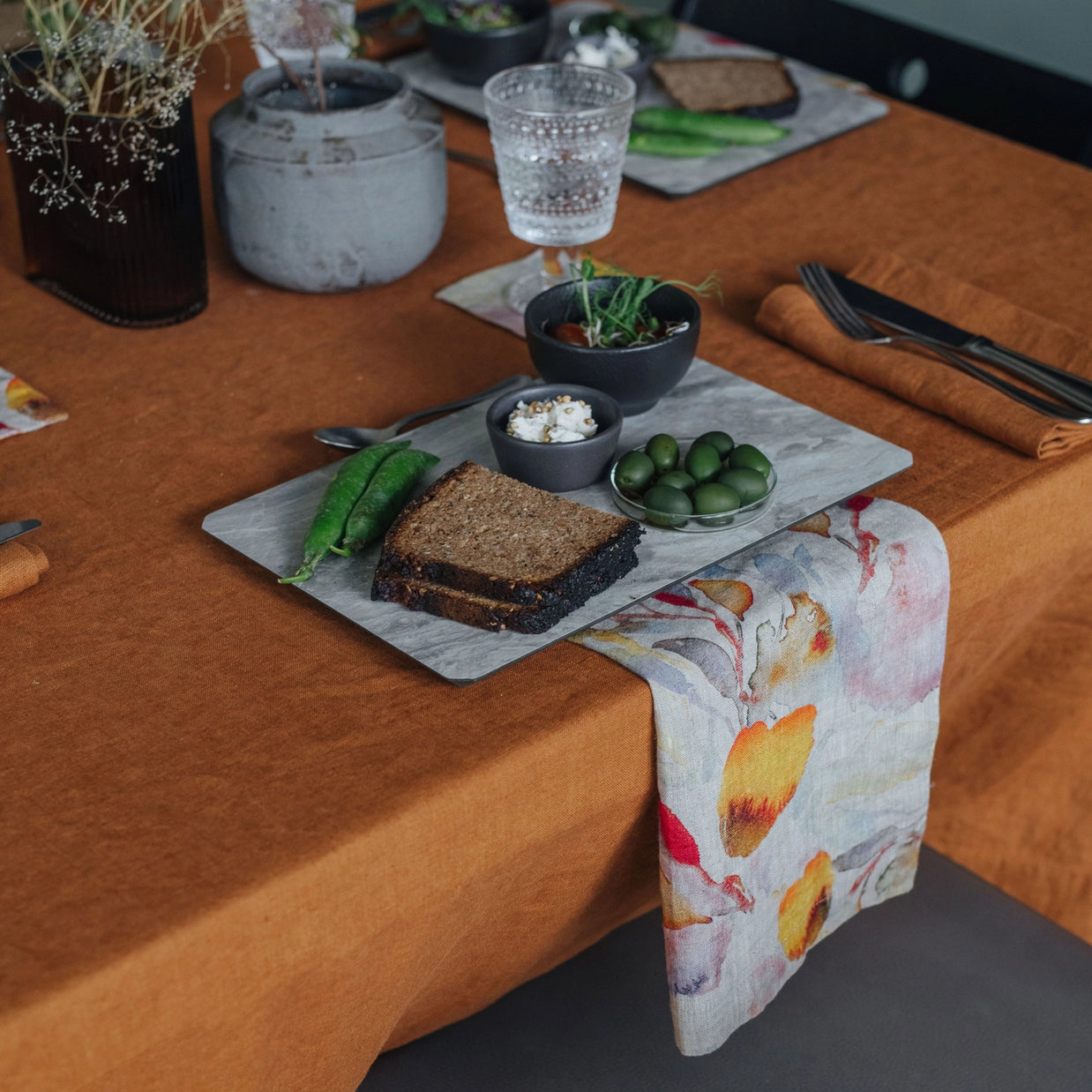 Linen table runner with watercolor botanical print in coral, yellow and gray tones draped across burnt orange tablecloth
