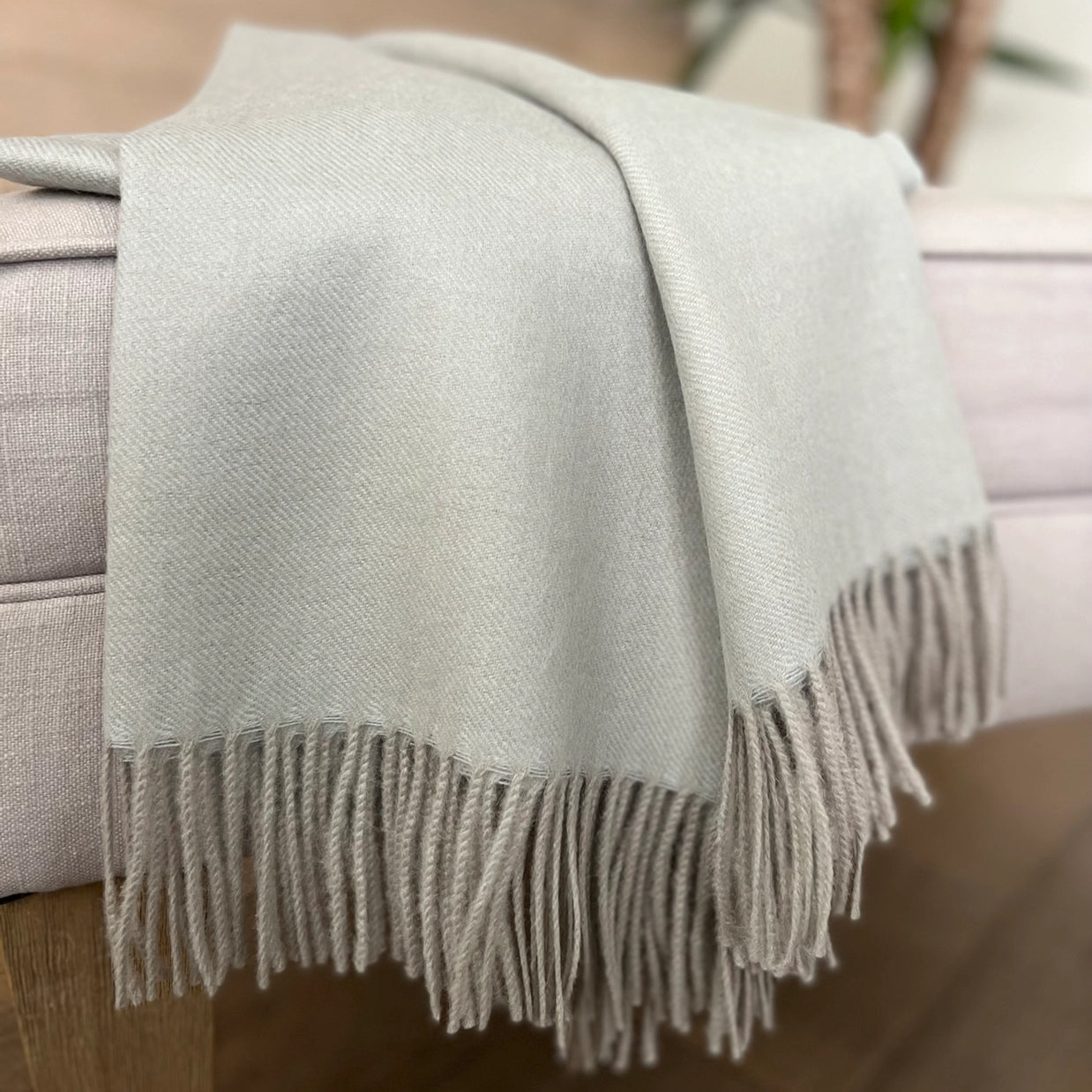 Pale sage green woven throw blanket with twisted fringe trim draped over beige linen sofa arm
