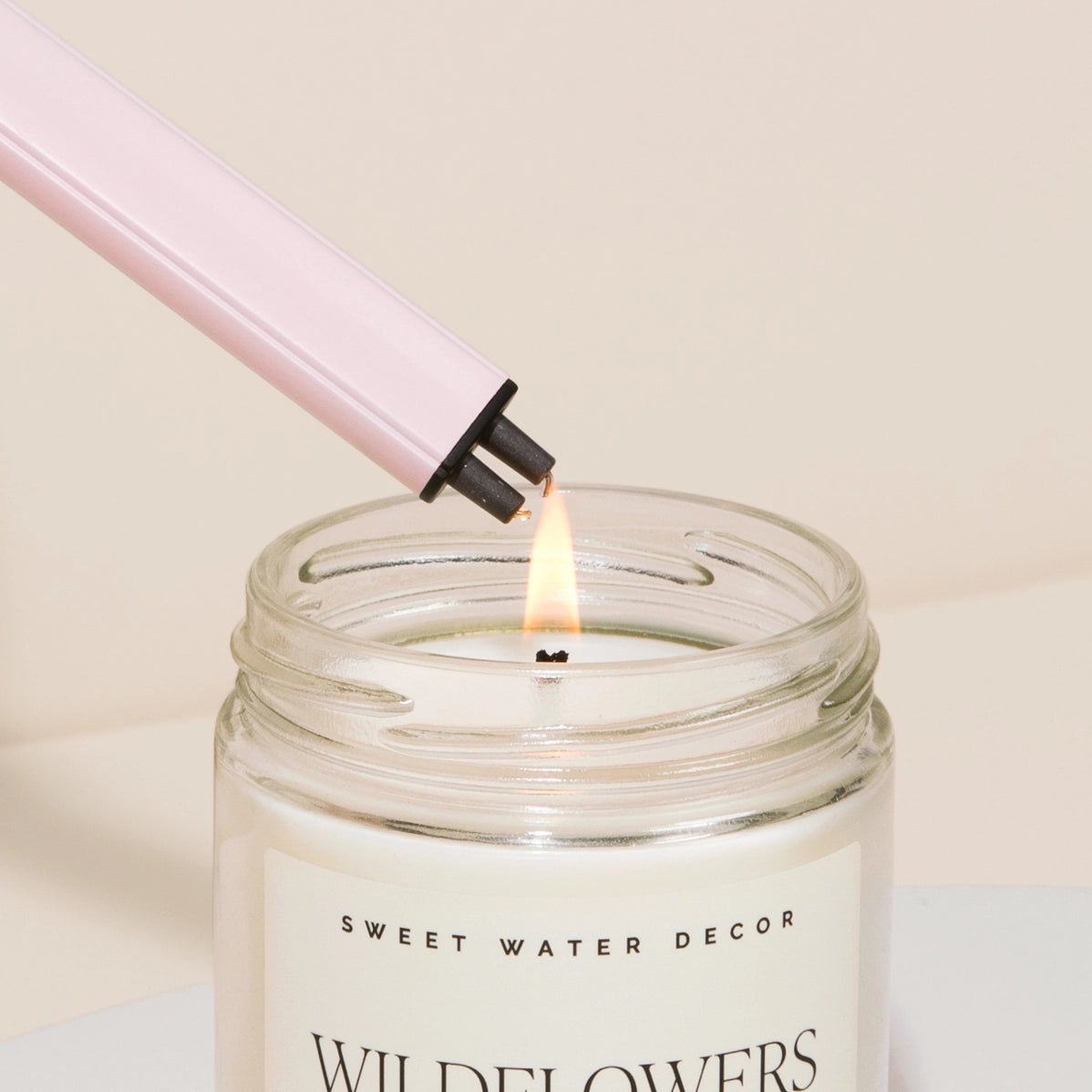 Pale pink candle lighter with black igniter touching lit wick in glass jar candle labeled Sweet Water Decor Wildflowers