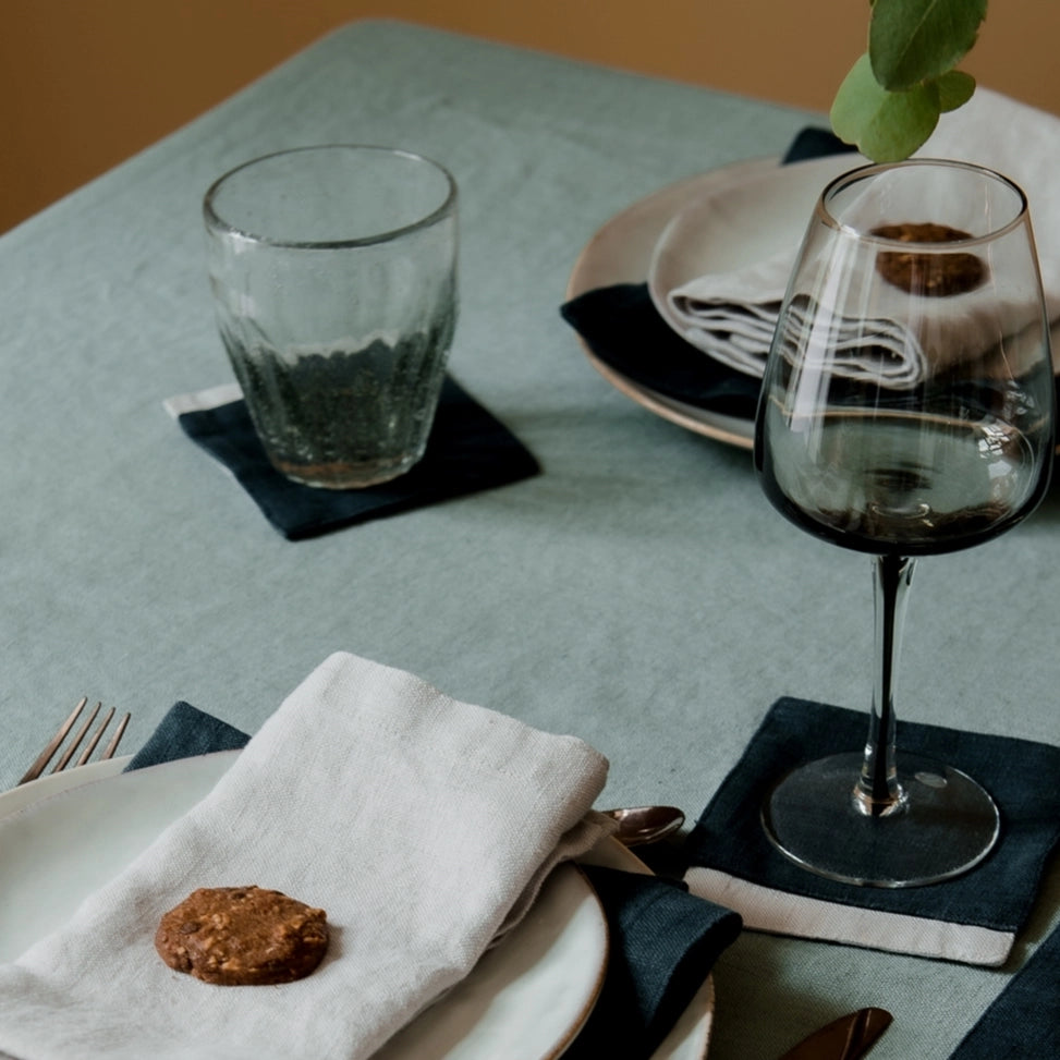 Slate blue linen tablecloth with place setting featuring smoked glass stemware, white napkin, and dark charcoal coasters