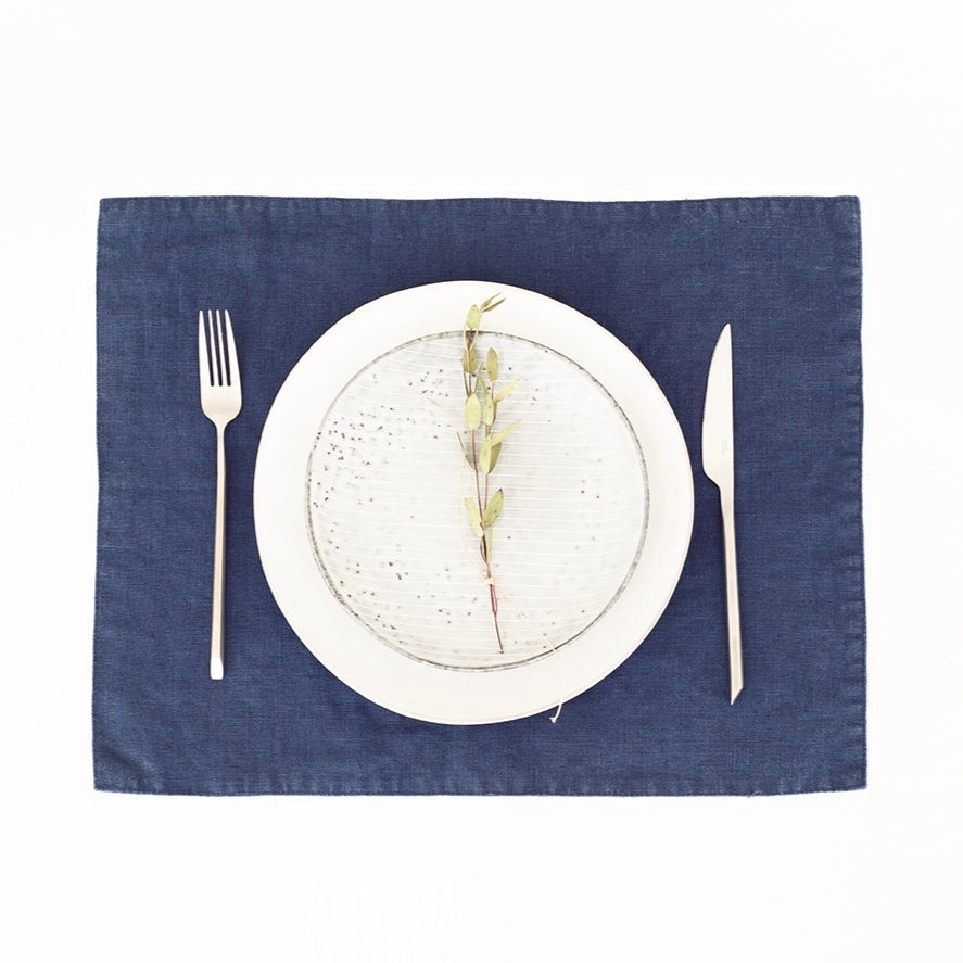 Navy blue linen placemat with textured weave, displayed with white dinnerware, silverware, and dried botanical stem