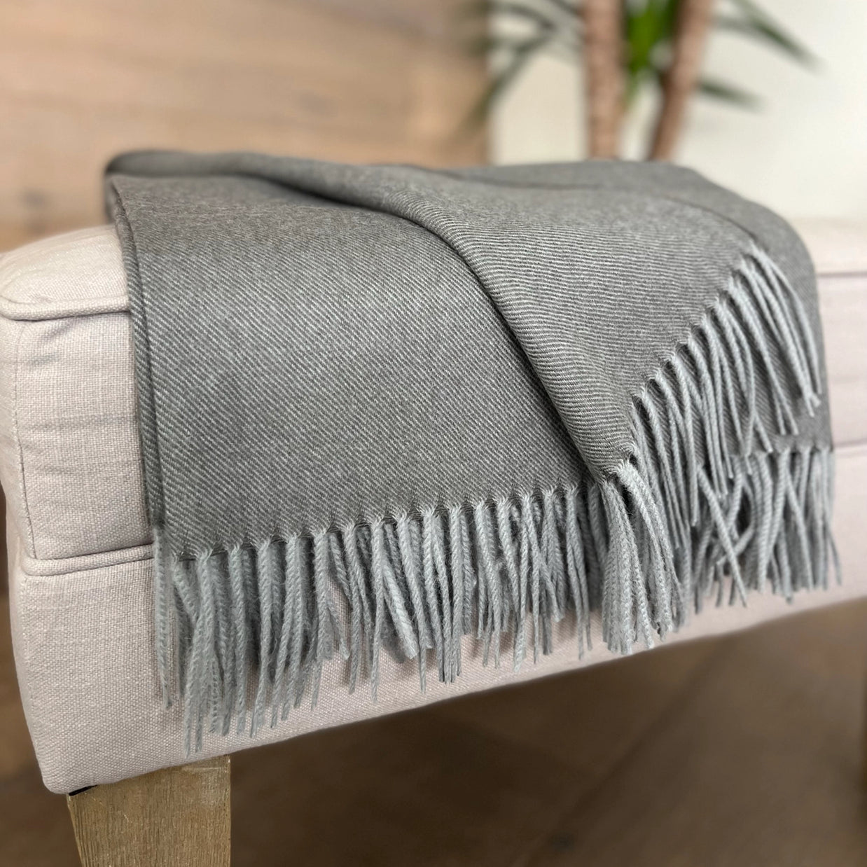 Charcoal gray wool throw blanket with diagonal twill weave and twisted fringe draped over beige linen ottoman