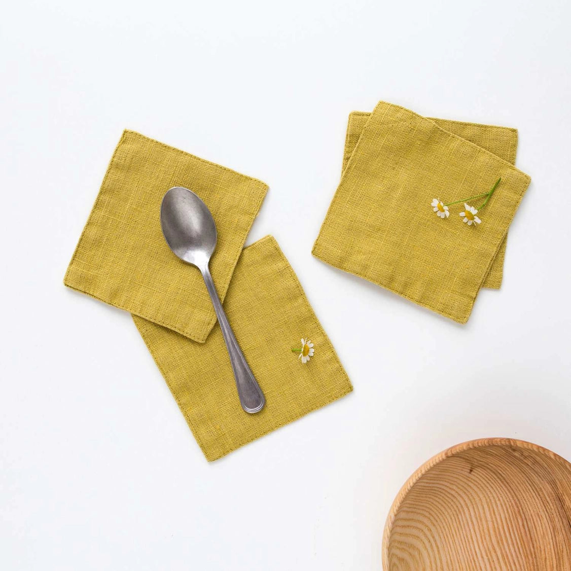 Mustard yellow textured linen coasters with small embroidered white daisy flowers, displayed with vintage silver spoon
