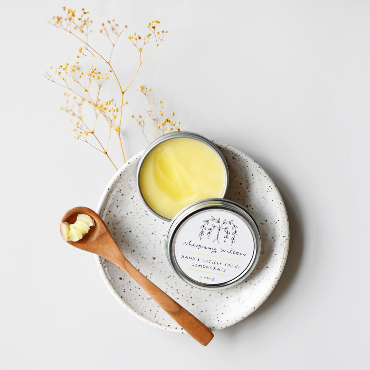 Lemongrass hand and cuticle salve in silver tin with yellow balm, wooden spoon, on speckled ceramic plate with dried flowers