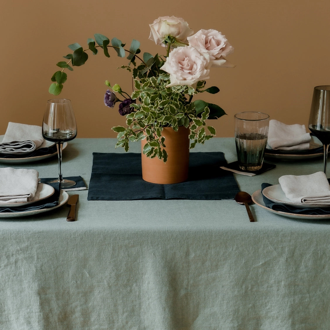 Sage green linen tablecloth with textured weave on dining table set with ceramic plates, wine glasses, and terracotta vase