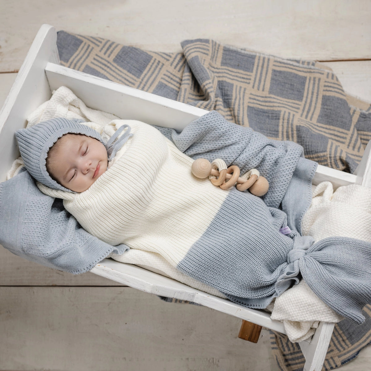 Newborn in gray knit bonnet and blue-cream colorblock sleep sack holding wooden teething rings in white wooden crate