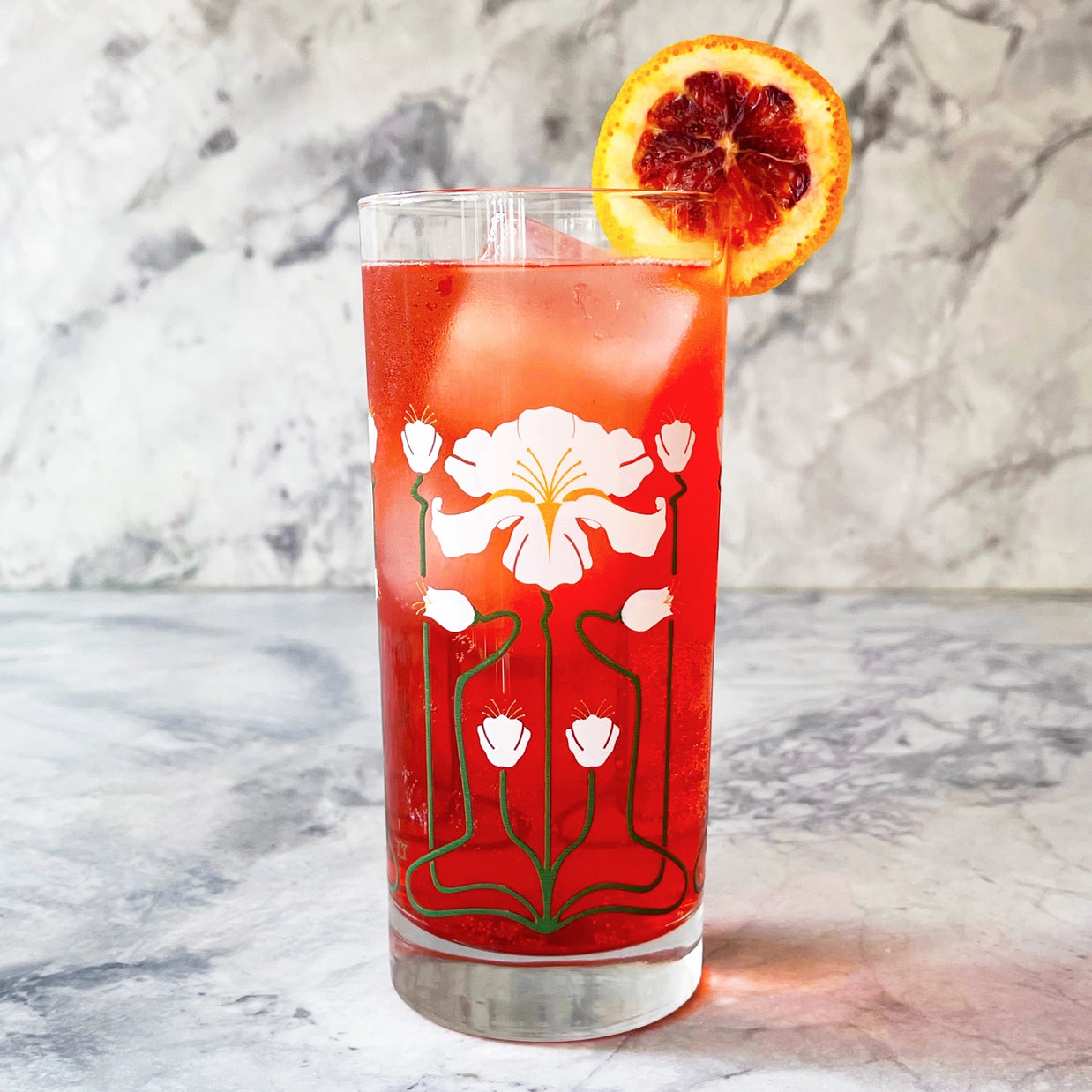 Tall glass with white art nouveau floral pattern filled with red cocktail, ice sphere, and blood orange slice garnish