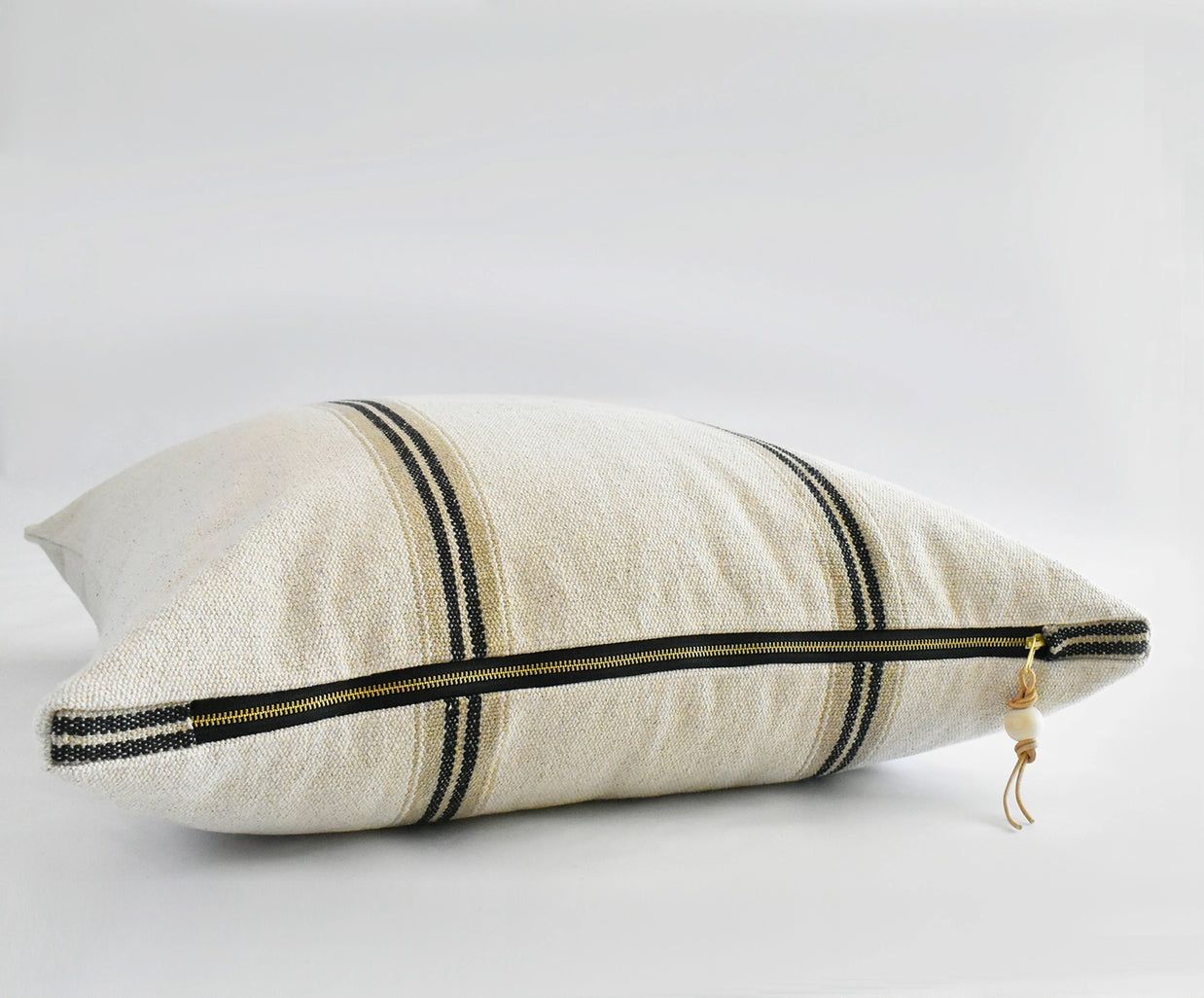Cream linen lumbar pillow with vertical black double stripes and brass zipper with leather pull tab on neutral backdrop