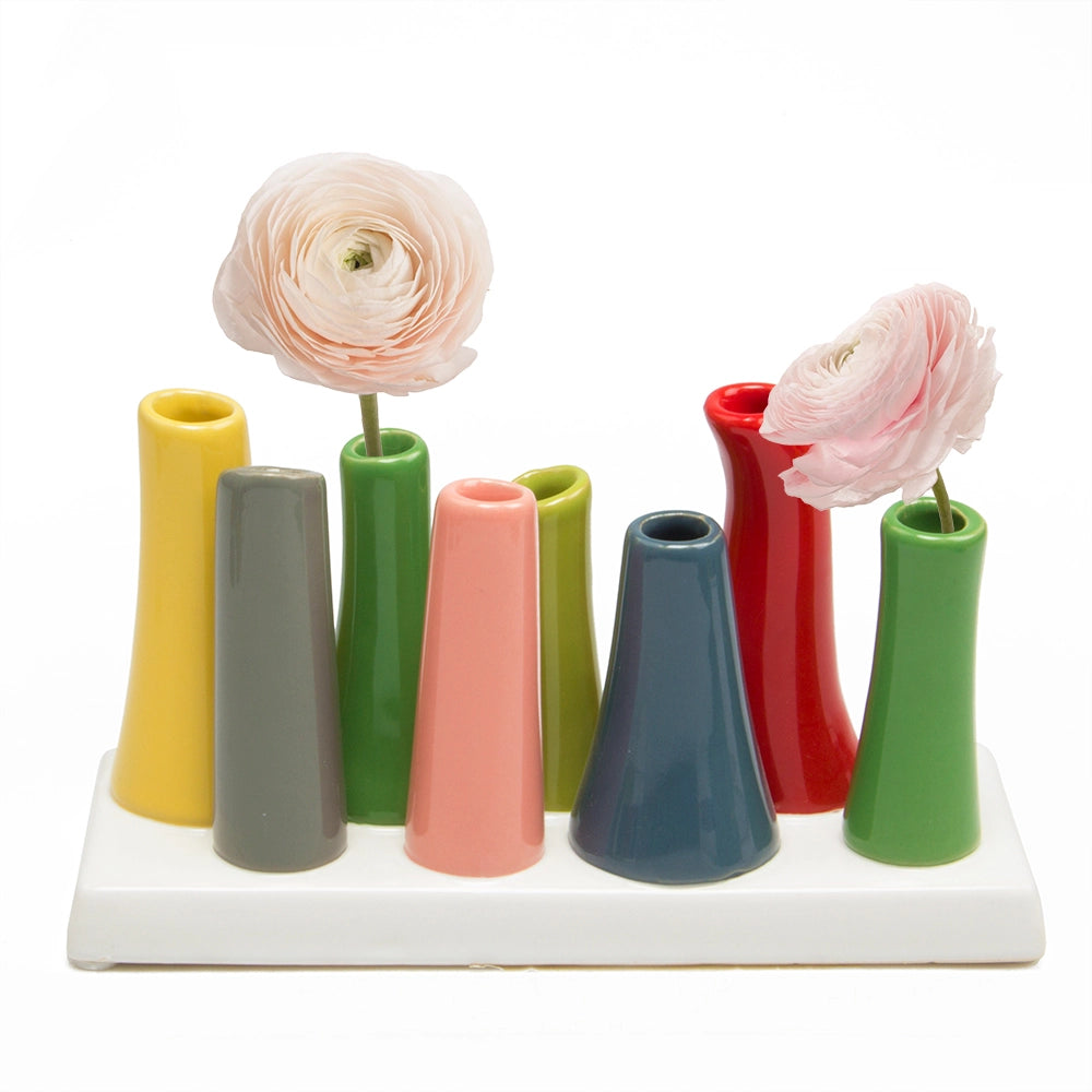 Ceramic bud vase set with nine colorful cone-shaped tubes on white tray holding two pale pink ranunculus flowers