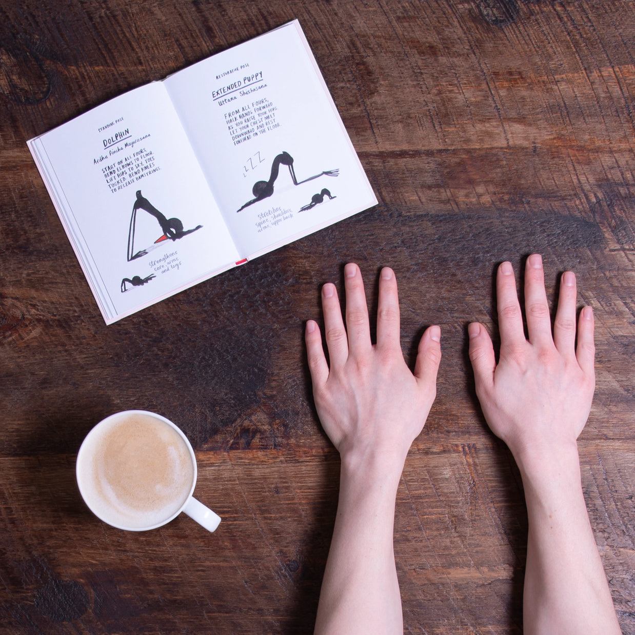 Illustrated yoga pose book open on rustic wooden table with hands and white coffee cup nearby, showing dolphin and extended puppy poses