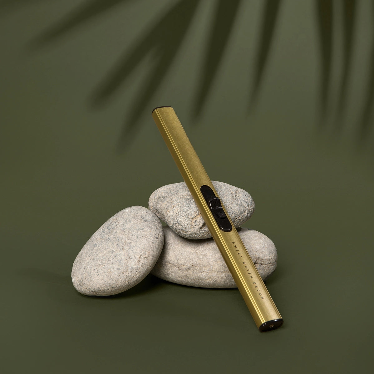 Brushed gold metal vape pen propped against stacked smooth gray river stones on sage green background with palm shadow