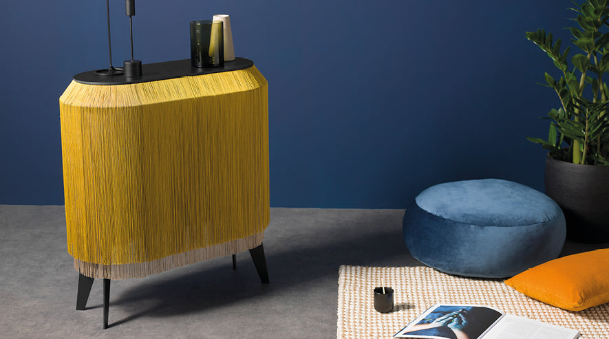 Hexagonal side table with golden yellow fringe wrapped around black top on angled legs beside blue velvet ottoman pouf