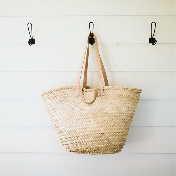 A French Basket Tote Bag with double handles, made from palm leaves, hanging on a white wall hook.