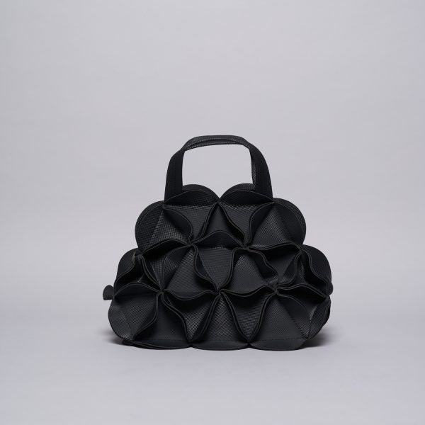 Black leather sculptural handbag with three-dimensional geometric petal-like panels and rounded top handle on gray background