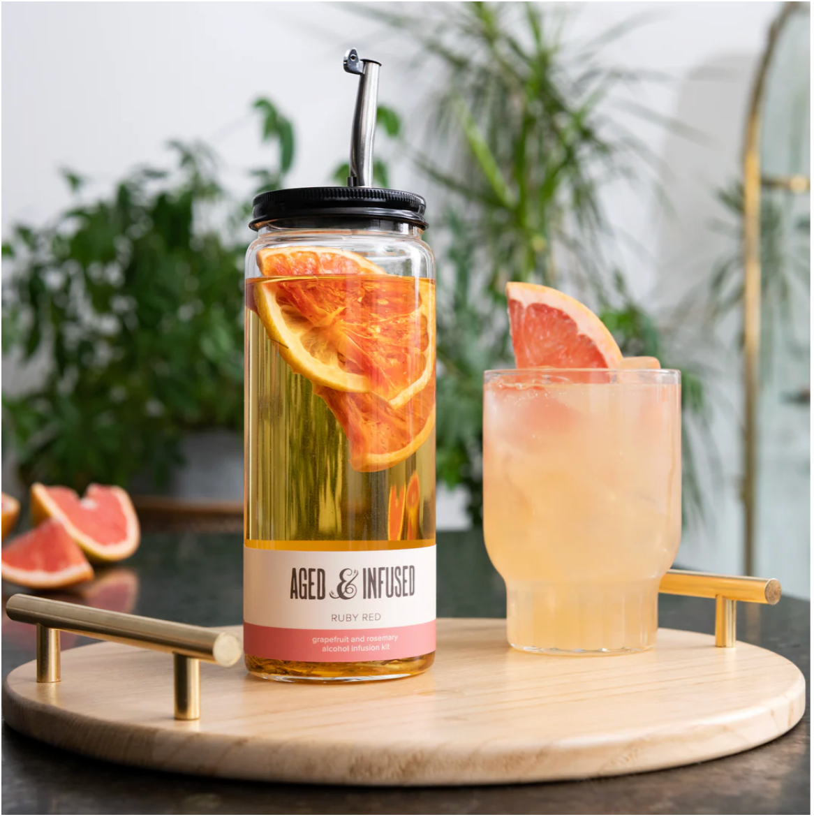 Glass infusion jar with black lid and metal straw containing grapefruit and rosemary in golden liquid beside frosted cocktail