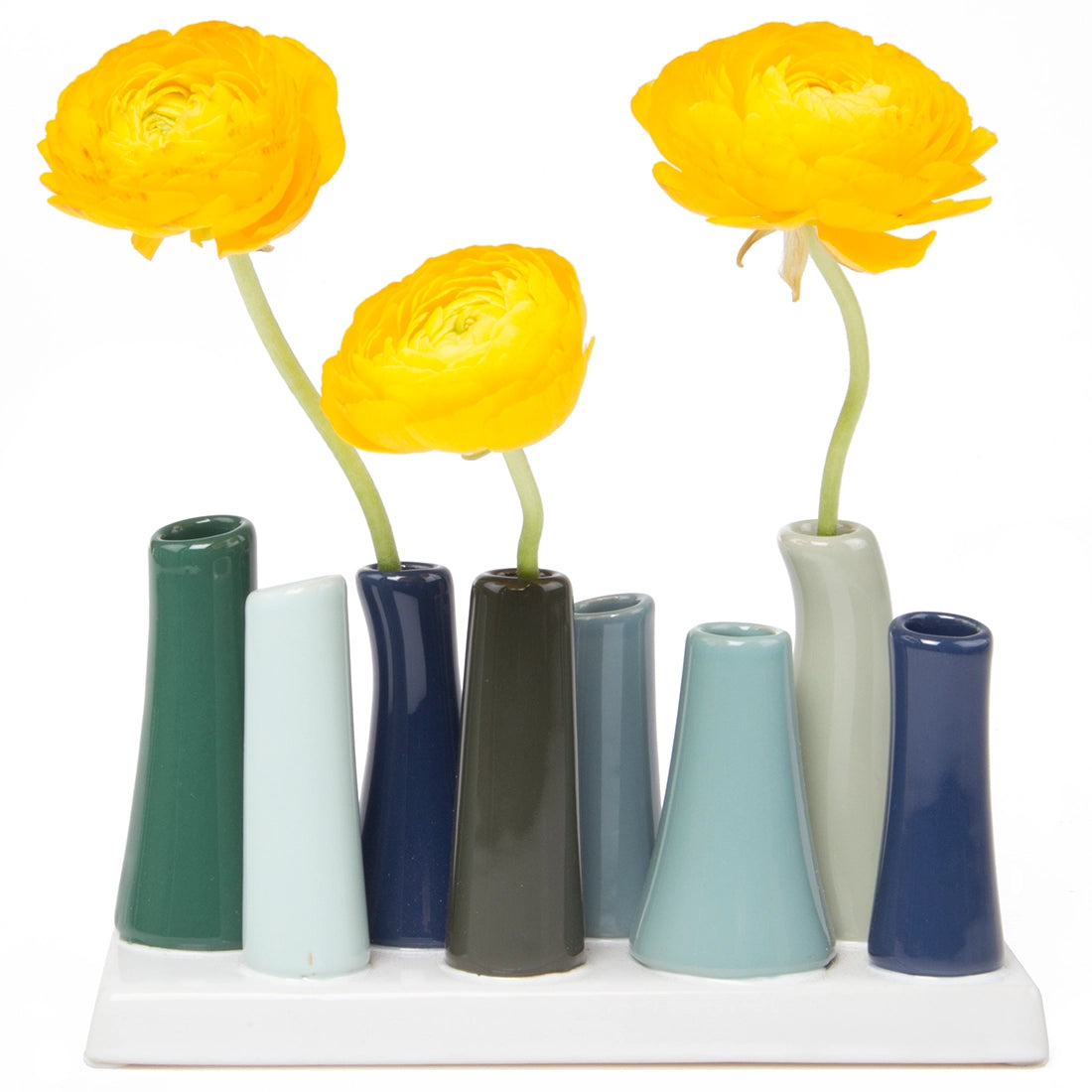 Graduated ceramic bud vase set with seven tapered tubes in blue, green, and sage tones holding bright yellow ranunculus