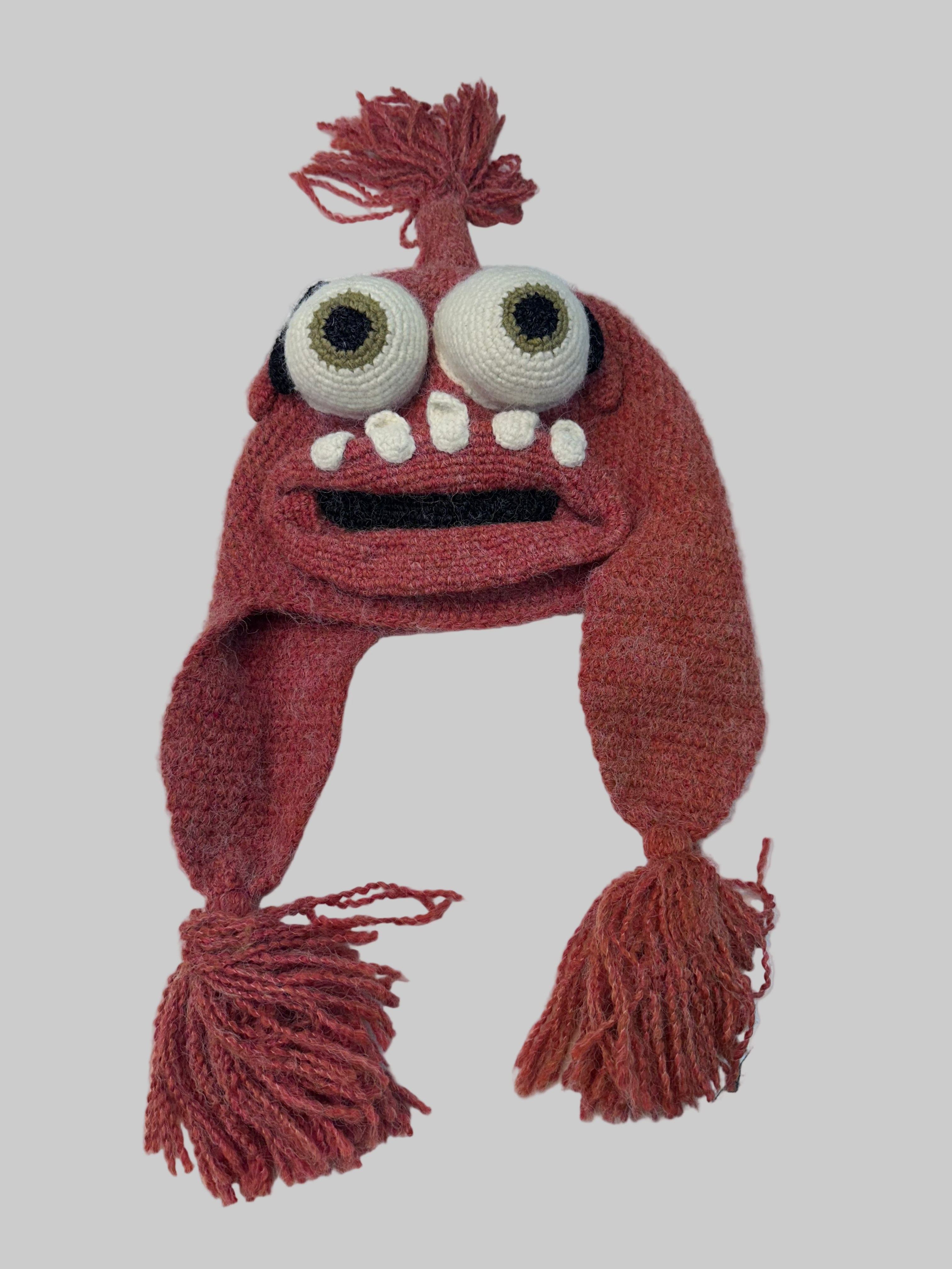 Hand-crocheted monster winter hat in dusty rose yarn with bulging white eyes, white teeth, braided earflaps and tassel trim