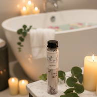 A clear cylindrical bottle of bath salts with a black cap and a white label reading 'The Inspiration Haven Bath Salts Calming' on a bathroom sink, with a luxurious tub and candles in the  background.