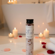 A clear cylindrical bottle of bath salts with a black cap and a white label reading 'The Inspiration Haven Bath Salts Self Love' sits on a bathroom floor in front of a luxurious bath tub and candles.