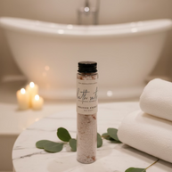 A clear cylindrical bottle of bath salts with a black cap and a white label reading 'The Inspiration Haven Bath Salts Positive Energy' sits on a marble table next to a inviting bath tub
