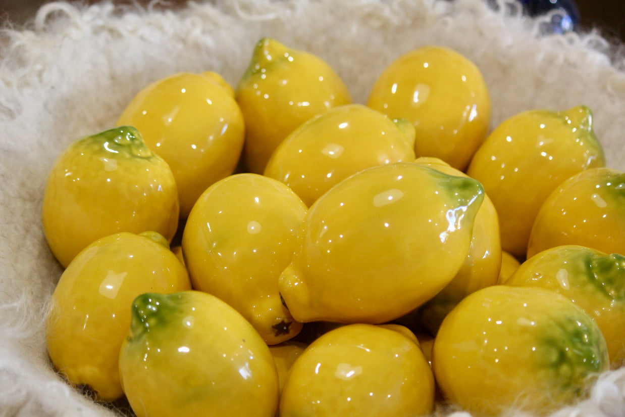 Glossy ceramic lemon replicas in bright yellow with green stems, nestled in cream woven basket with realistic texture