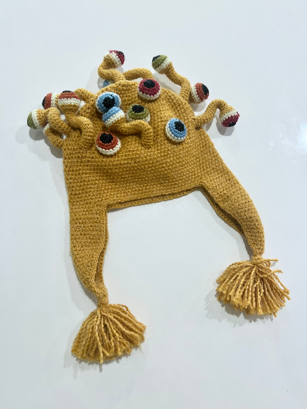 Mustard yellow crocheted monster hat with multiple eyeball tentacles in blue, green, and burgundy, braided ear flaps with tassels