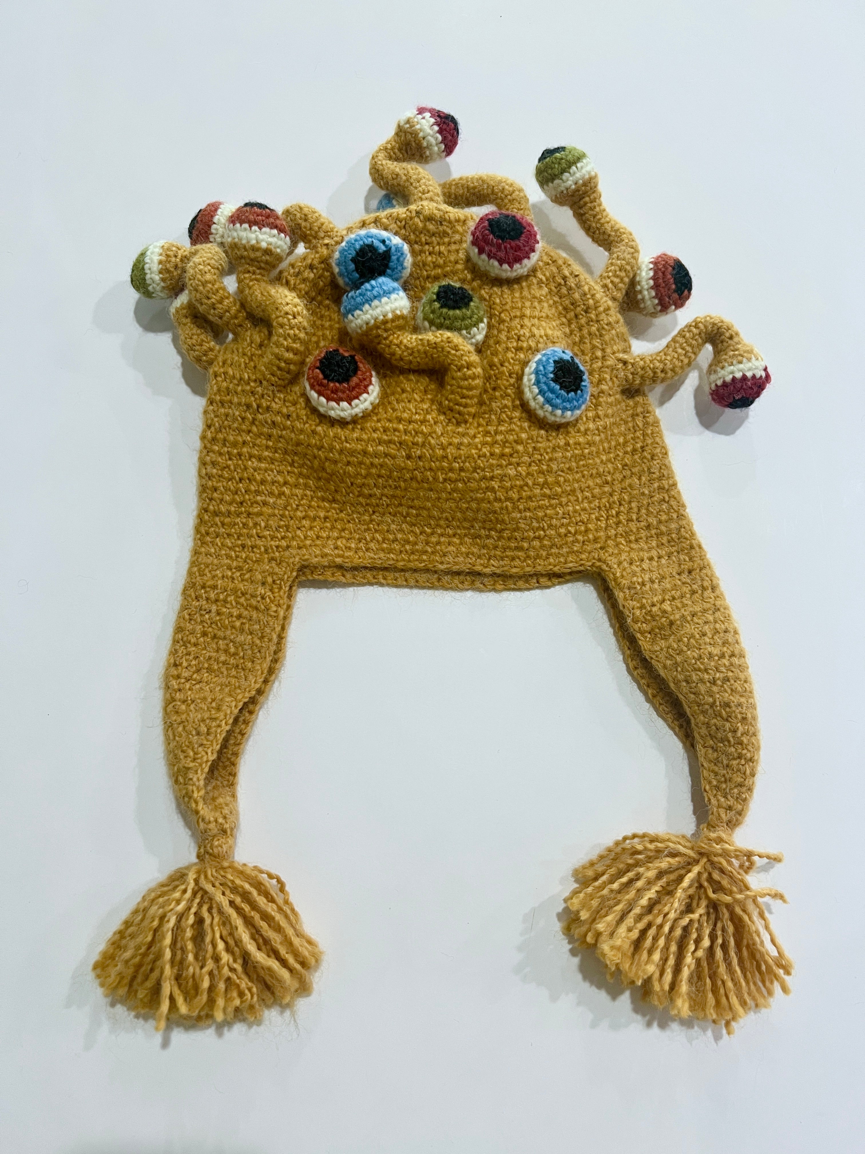 Mustard yellow crocheted winter hat with ear flaps, yarn tassels, and multiple colorful eyeball tentacles on top