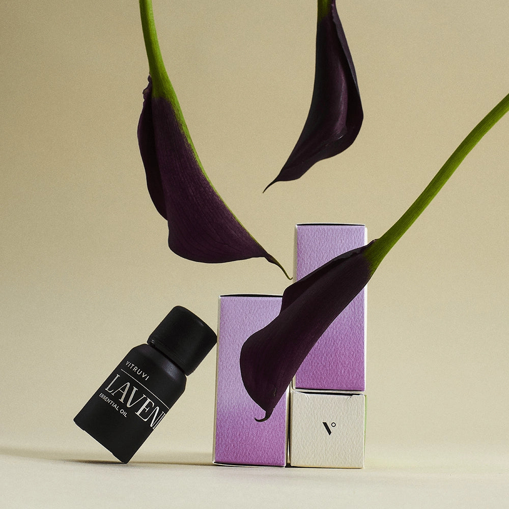 Black glass essential oil bottle with lavender label beside purple and cream boxes with dark purple calla lily stems