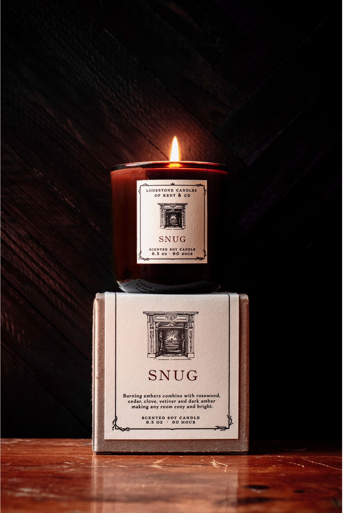Amber glass soy candle burning with cream label featuring fireplace illustration, displayed on matching gift box