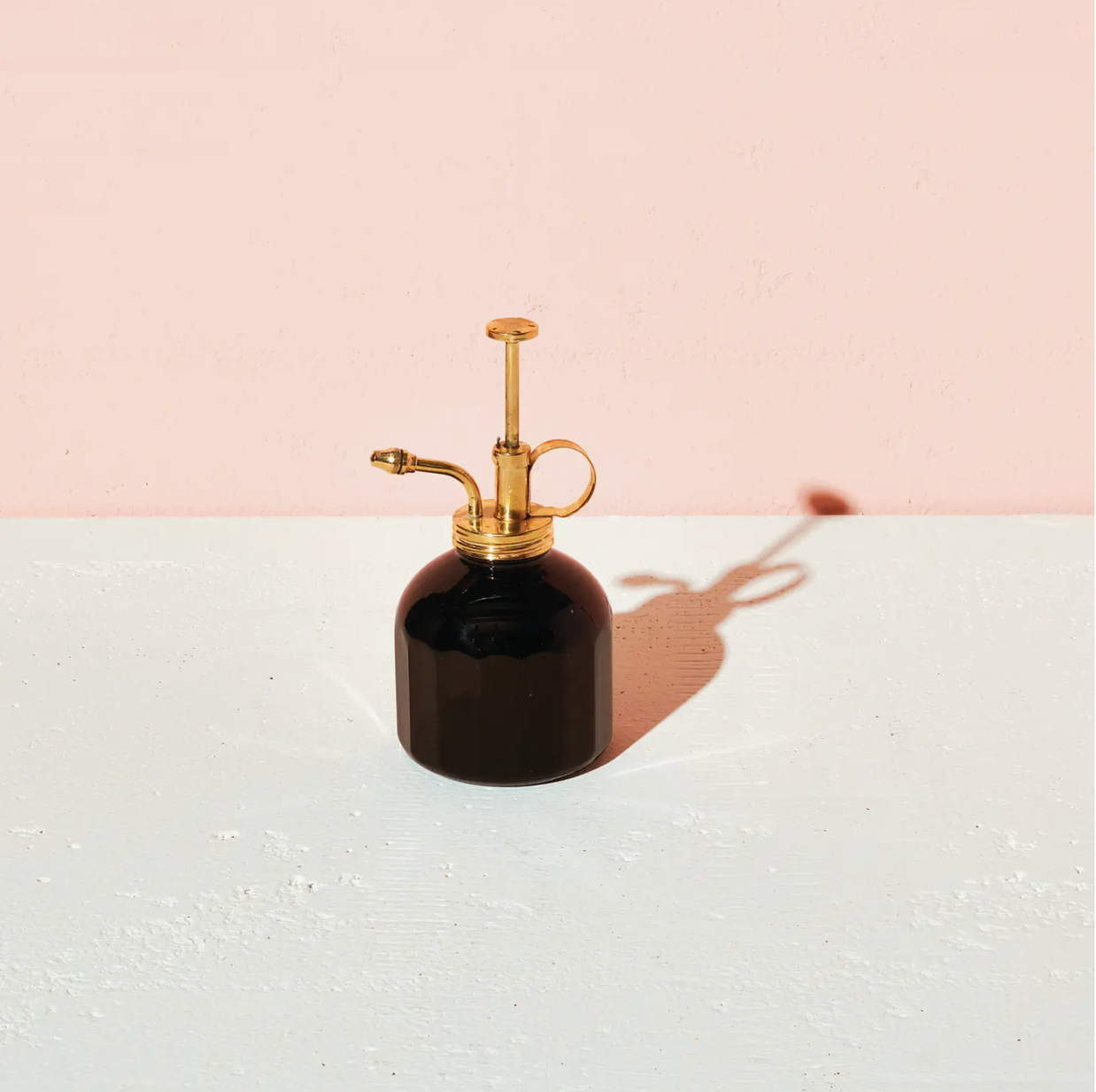Black glass plant mister bottle with brass pump sprayer and curved handle on white surface against pink background
