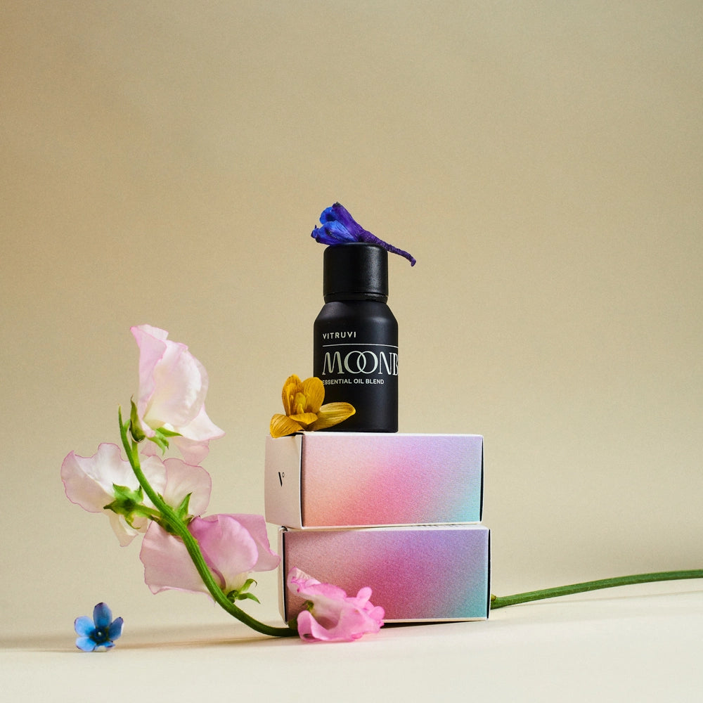 Black essential oil bottle labeled MOON VI atop gradient pink-blue boxes with fresh pink sweet peas and dried flowers