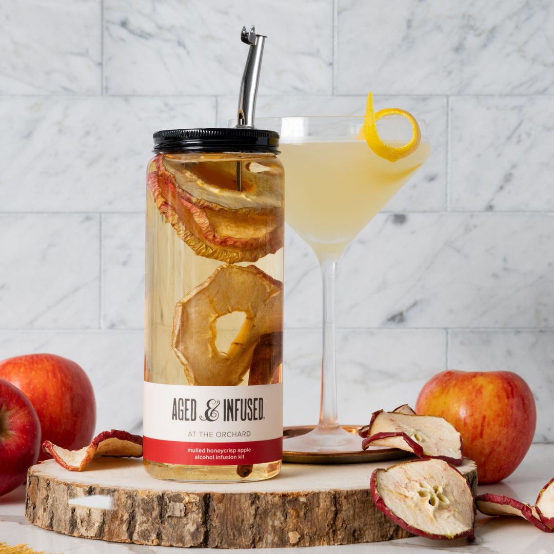 Glass jar with dried honeycrisp apple slices and metal spout beside pale cocktail with yellow lemon twist garnish
