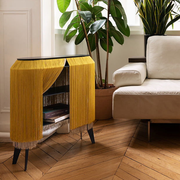 Mid-century modern side table with golden yellow fringe curtain doors, black tapered legs, and dark wood top on herringbone floor