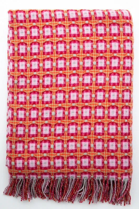 Woven throw blanket with repeating geometric pattern in pink, red, orange and white with twisted fringe trim along bottom edge