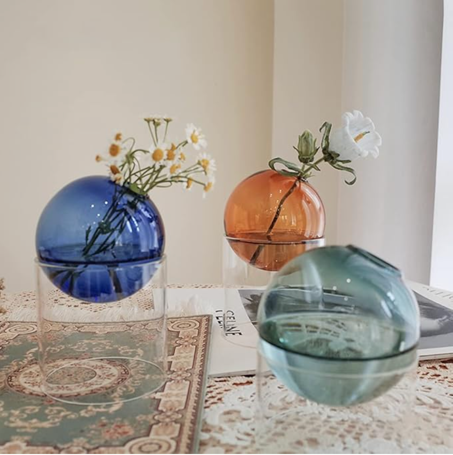 Three blown glass spherical vases in cobalt blue, amber orange, and sage green with clear glass bases holding delicate flowers