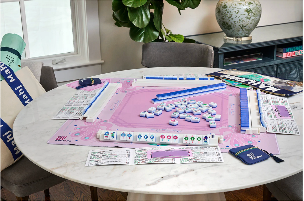 Mahjong game set on pink silicone mat with white tiles, blue tile racks, and navy blue card holders on marble table
