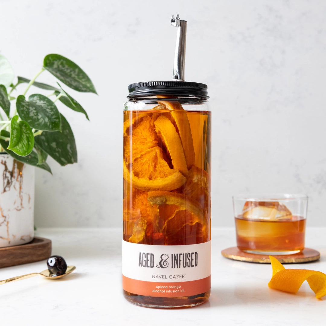 Glass jar with black lid and metal straw filled with amber liquid and dried orange slices labeled spiced orange infusion kit