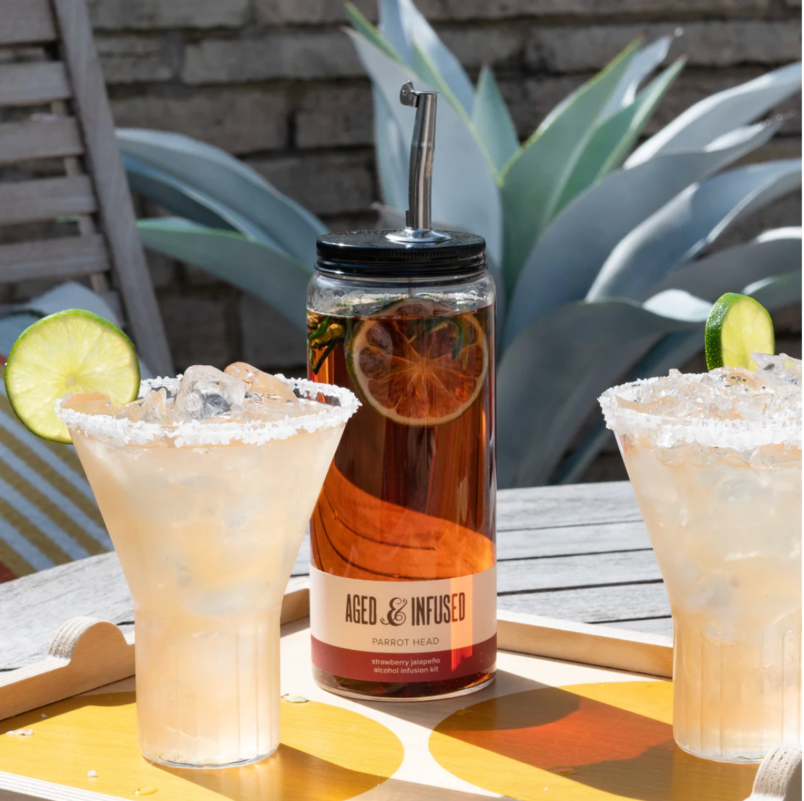Glass jar cocktail infusion kit with metal spout between two frosted margaritas with salted rims and lime garnish