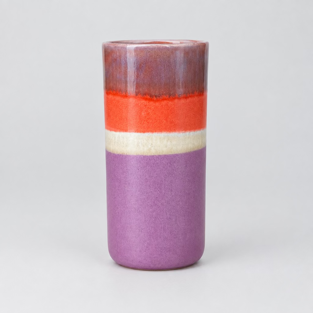 A handmade ceramic vase with a three-color glazed design, featuring a purple base, a cream band, and a bright orange-red top section, sitting on a light wood table in a pottery shop.