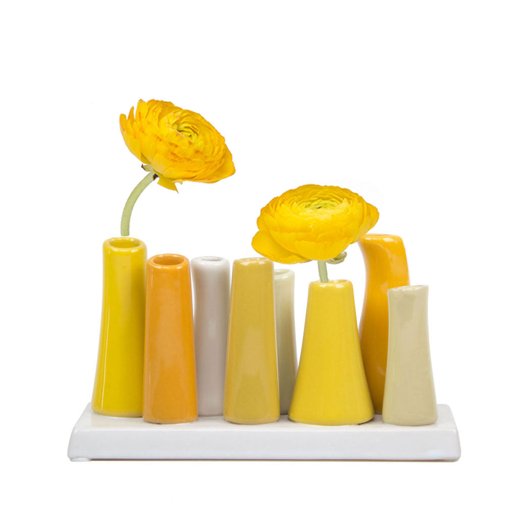 Eight ceramic tube vases in yellow, orange, and cream tones on white tray with two bright yellow ranunculus flowers