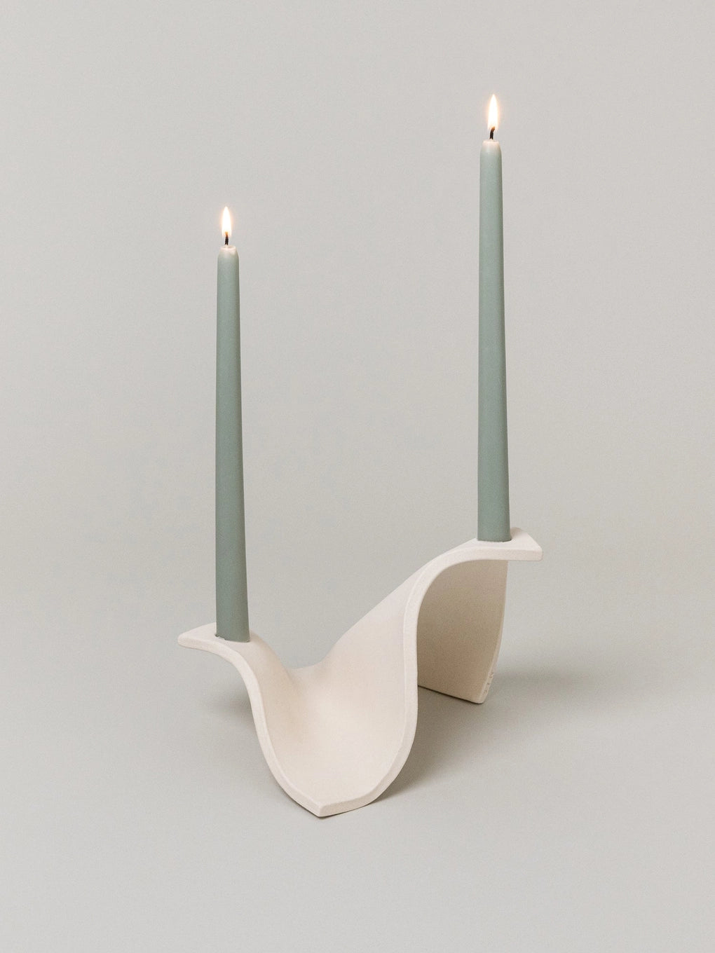 Modern white ceramic candelabra with curved sculptural design holding two tall sage green taper candles with lit flames
