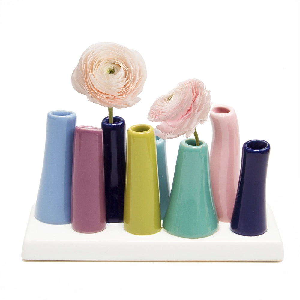 Multi-tube ceramic bud vase with eight conical openings in pastel colors on white base, two holding pale pink ranunculus