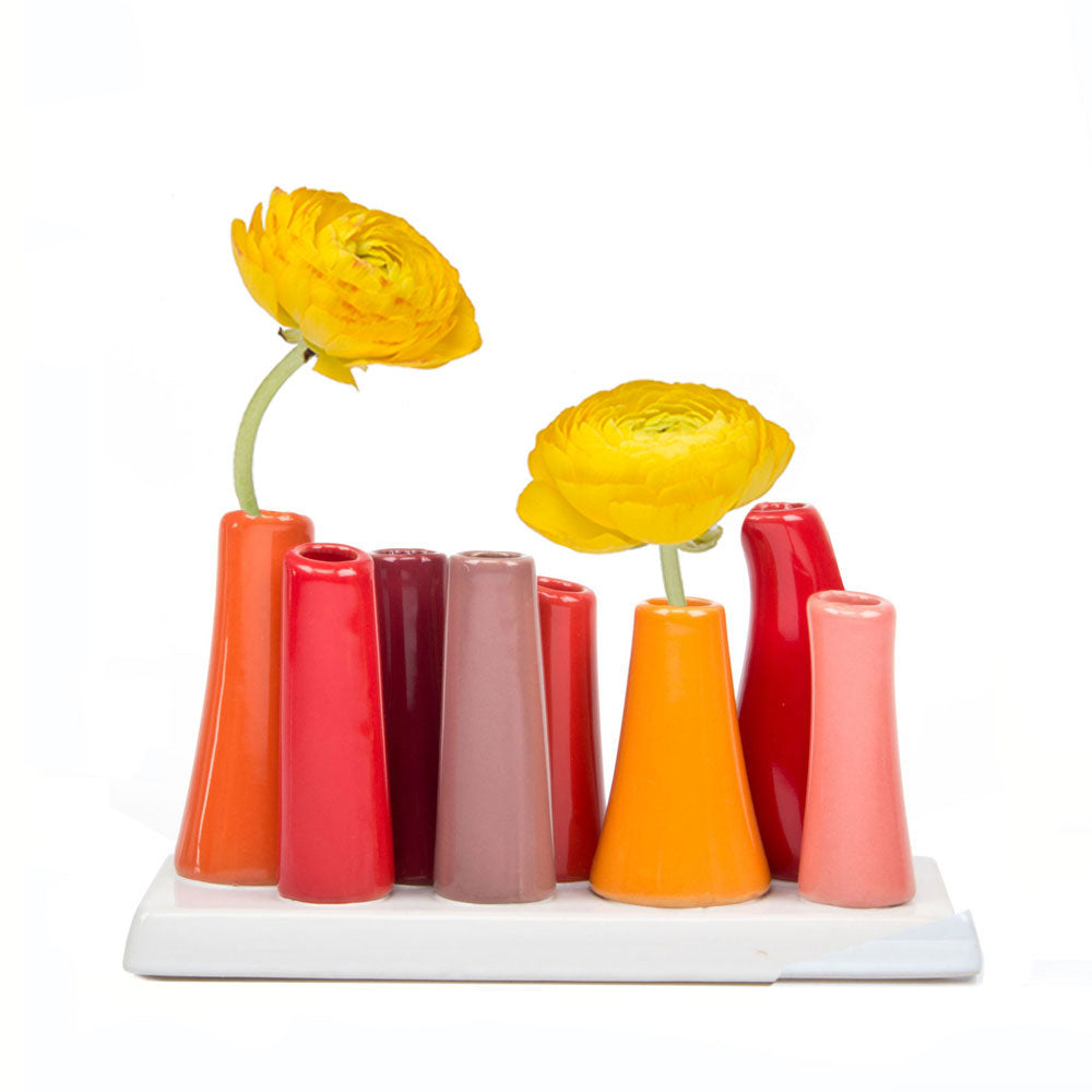 Ceramic bud vase set with eight tapered tubes in coral, pink, burgundy, and orange shades holding two yellow ranunculus flowers